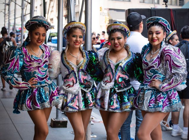 Four women in colorful, sequined costumes pose in a vibrant street parade in New York City
