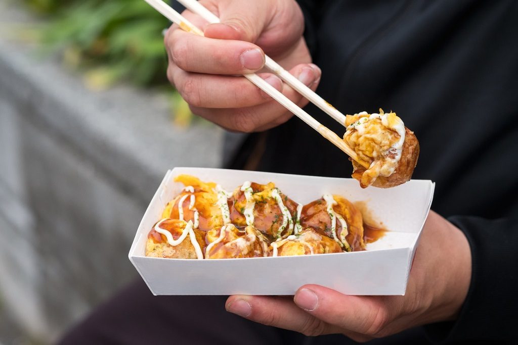 Kyoto man eating traditional Japanese cuisine - takoyaki balls from a take out container while people watching on Cat Street (Kyu Shibuya-gawa Promenade) near Harajuku Street, in Tokyo, Japan. The takoyaki is covered with mayonnaise and teriyaki sauce.