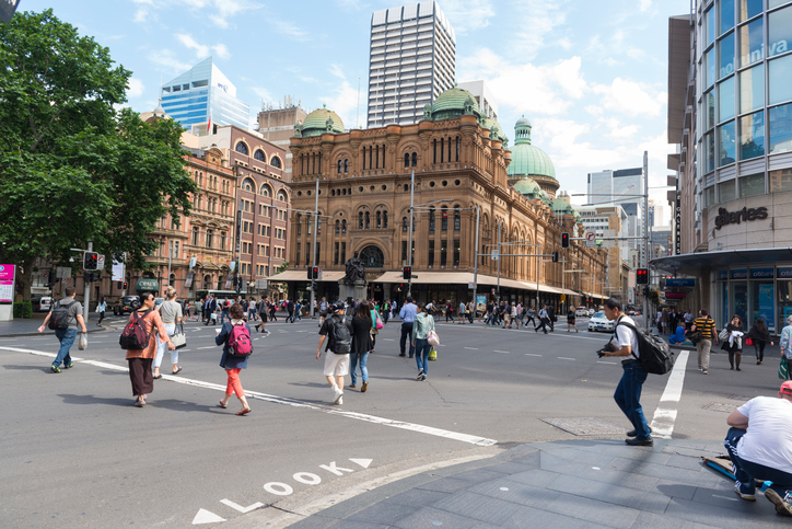 People wait on the corner of George Street and Park Street, opposite of Queen Victoria Building while vehicles pass by on Market Street.