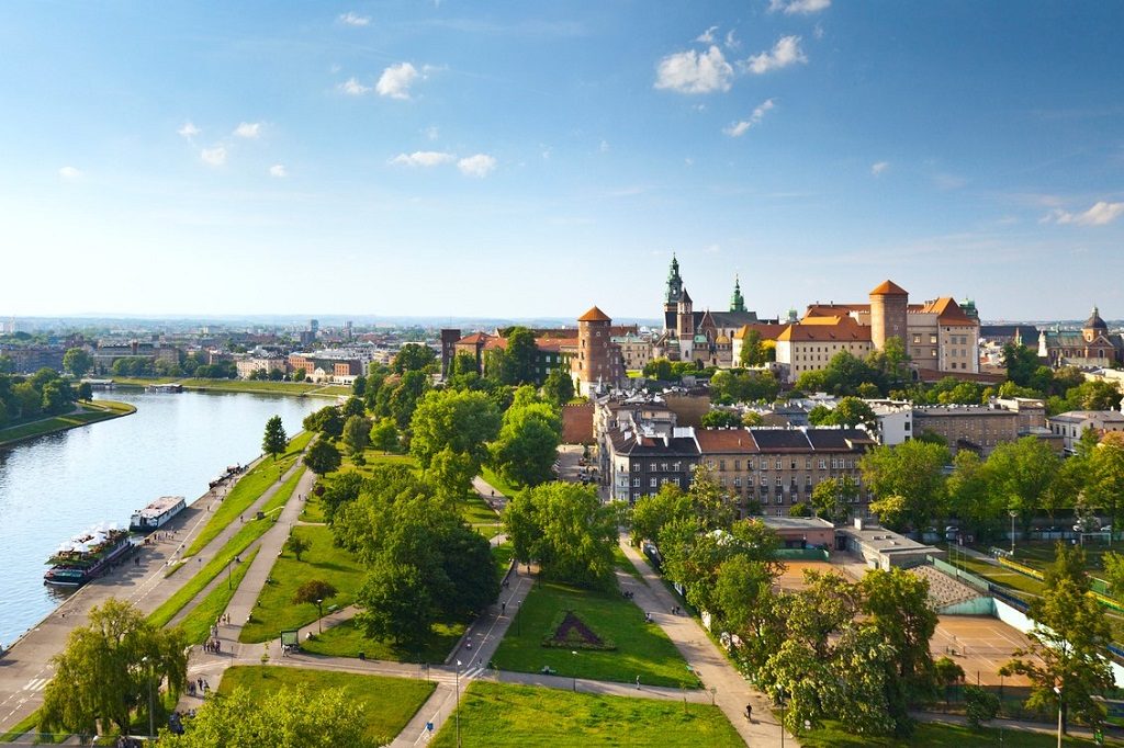 Daytime panorama of Krakow, Poland, featuring Wawel Castle. The Vistula River flows along the left side of the image, and the castle is visible on the right, near the horizon. There is a blue sky with sparse, wispy clouds above the horizon and a manicured lawn intersected by walking paths in the foreground.