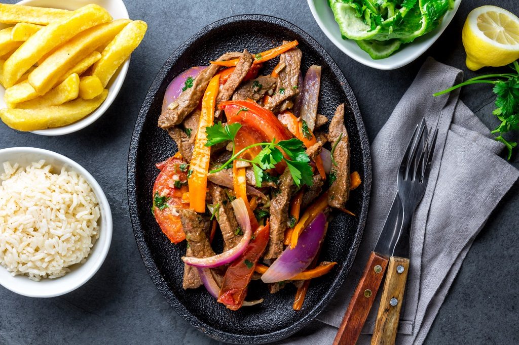 Peruvian dish Lomo saltado - beef tenderloin with purple onion, yellow chili, tomatoes served on black plate with french fries and rice. Top view