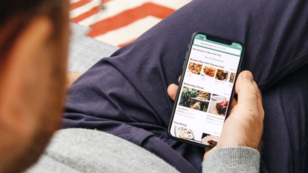 Paris: Man holding using the Tripadvisor hotel booking app on the new Apple iPhone X 10 Display while seating in couch with Retina Display screen featuring all home apps