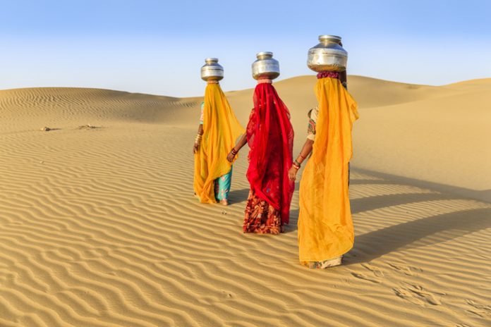 Indian women crossing sand dunes and carrying on their heads water from local well, Thar Desert, Rajasthan, India. Rajasthani women and children often walk long distances through the desert to bring back jugs of water that they carry on their heads.