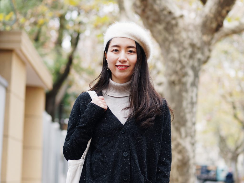 Beautiful young brunette woman in white beret and black woolen sweater smiling at camera with blur street background. Outdoor fashion portrait of glamour young Chinese cheerful stylish lady.