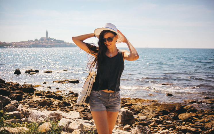 Vintage toned portrait of a young fashionable brunette relaxing by the sea in Rovigno, Istria, Croatia. She is resting in the hot summer day, wearing a dark grey top, jeans shorts, carrying a tote bag and a hat, walking by the sea. Shot in letterbox image ratio for more horizontal copy space with film emulation color grading.