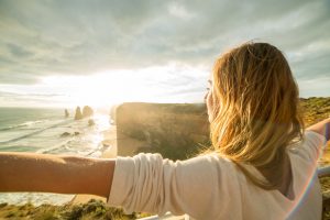 Cheering young woman arms outstretched at sunset. Shot at the Twelve Apostles sea rocks on the Great Ocean Road in Victoria's state of Australia.