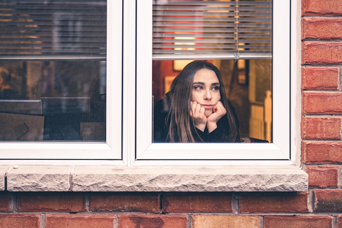 Young woman sitting inside her home, close to the window looking out. She is practicing social distancing due to pandemic of COVID-19 and stay at home order.