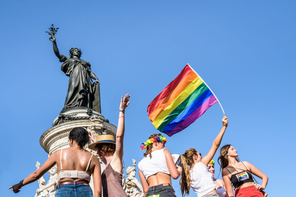 Six young women, one waving a rainbow flag high, stand in the sun at the foot of the statue of Marianne on the place de la Republique, where the Gay Pride parade ended.